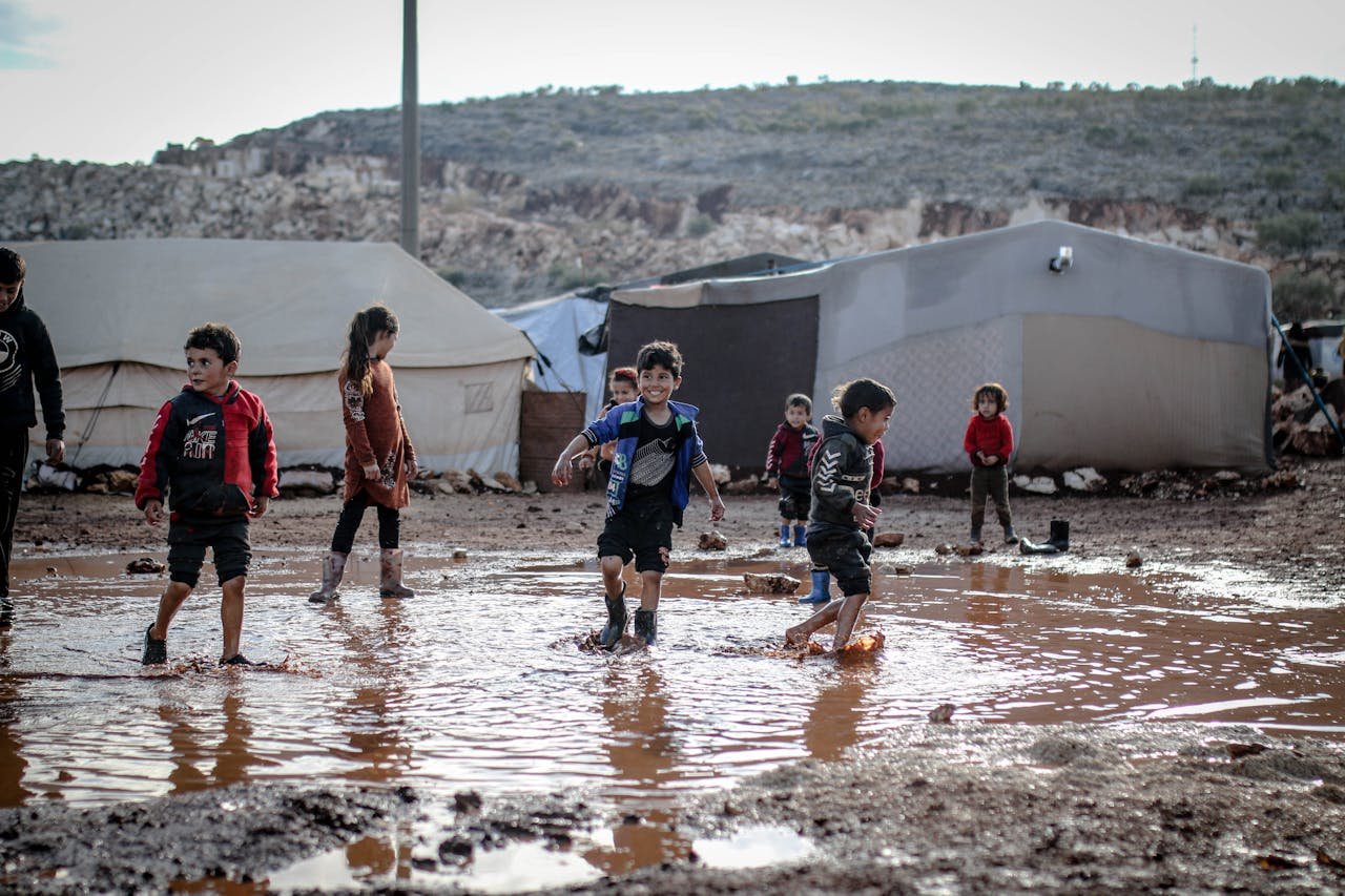 about-02 Kids enjoying playtime in a muddy refugee camp in Idlib, Syria amidst challenging conditions.